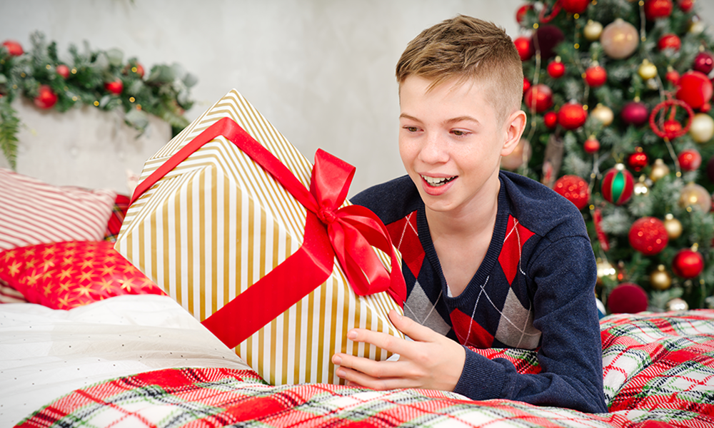 A cheerful teenager boy opens a Christmas gift. Cheerful Teenager lies in bed with a Christmas present in his hands